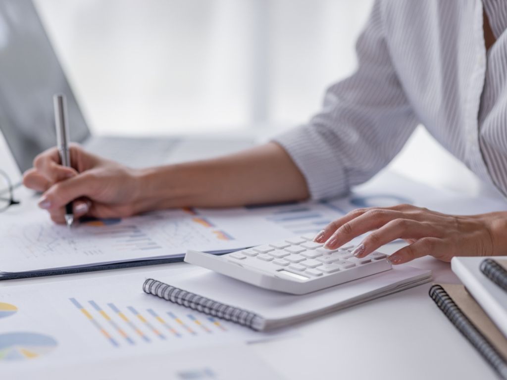 Una mujer en un escritorio, que no tiene un sistema de firma electrónica, utiliza una calculadora y escribe en papeles con la mano