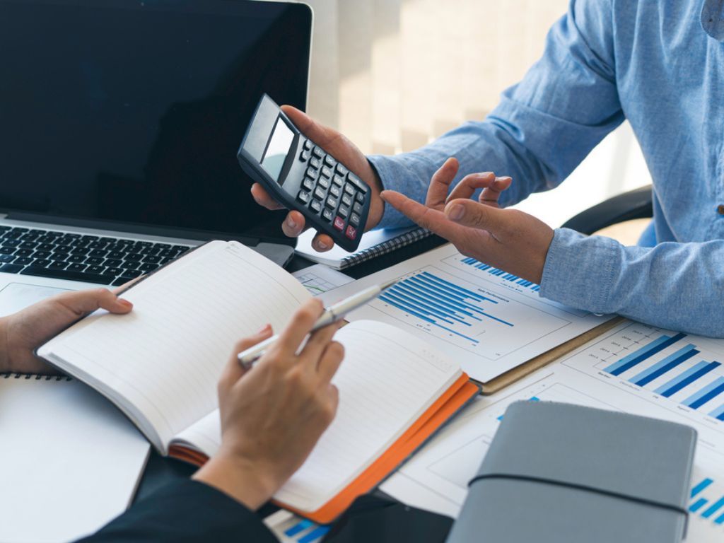 Dos pares de manos trabajan en la contabilidad para Pymes arriba de un escritorio lleno de documentos con gráficos. Una persona sostiene una calculadora, y la otra un lápiz y libreta.