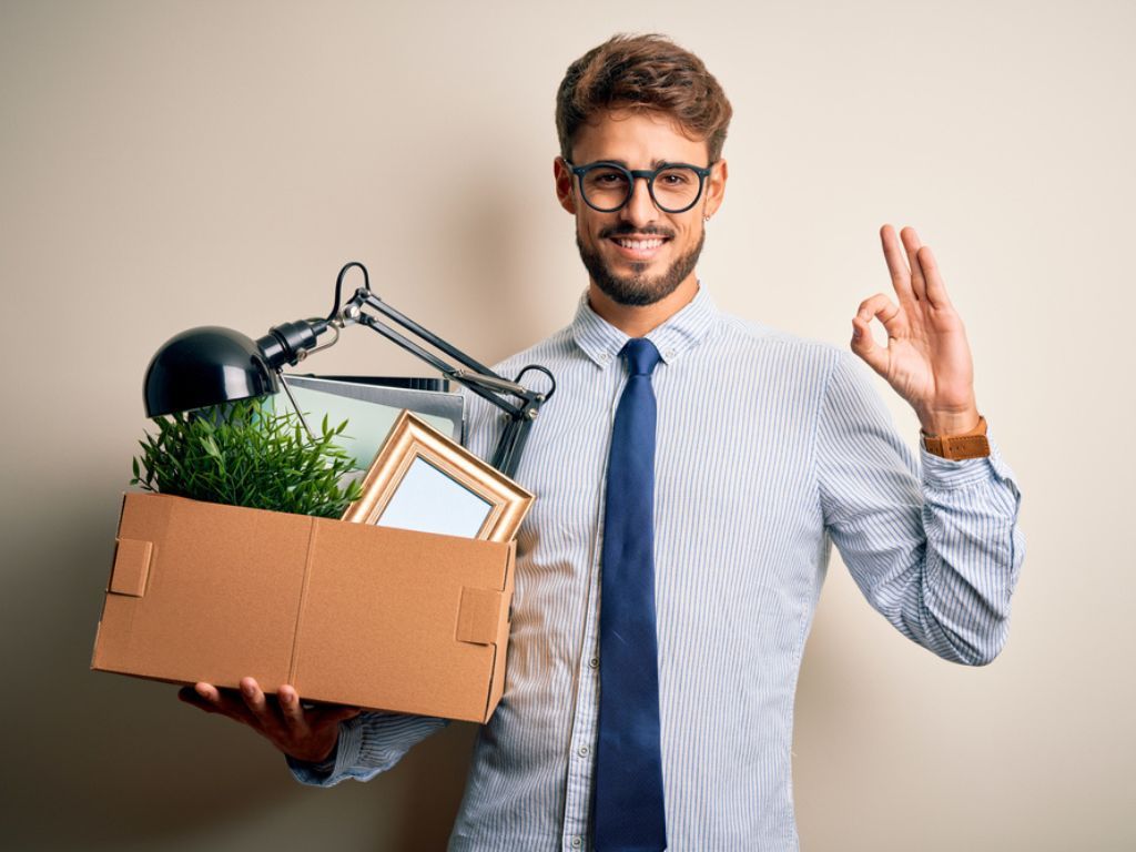 Un hombre sostiene una caja con sus pertenencias después de ser despedido pero sonríe aliviado al descubrir lo sencillo que es firmar un finiquito online.