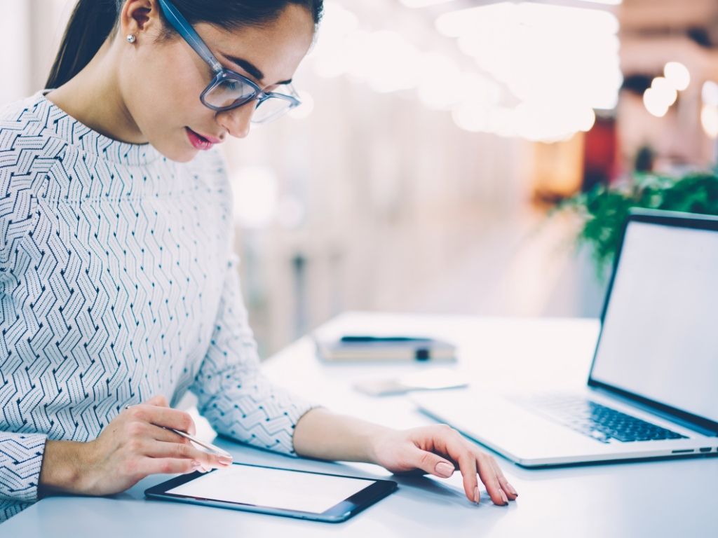 Mujer joven interactuando con una tableta en una oficina moderna, con una computadora abierta sobre la mesa.